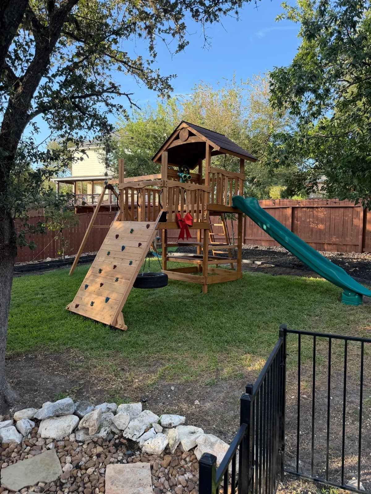 Wooden playset with a green slide, climbing ramp and tire swing in a sloping backyard with mature trees